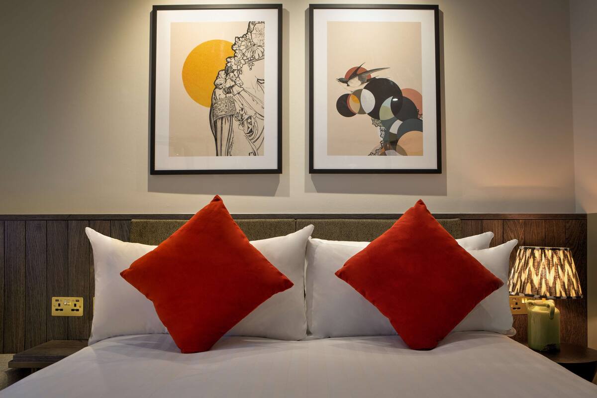 A hotel room with a bed, two red pillows, a nightstand with a lamp, and two framed artworks on the wall.