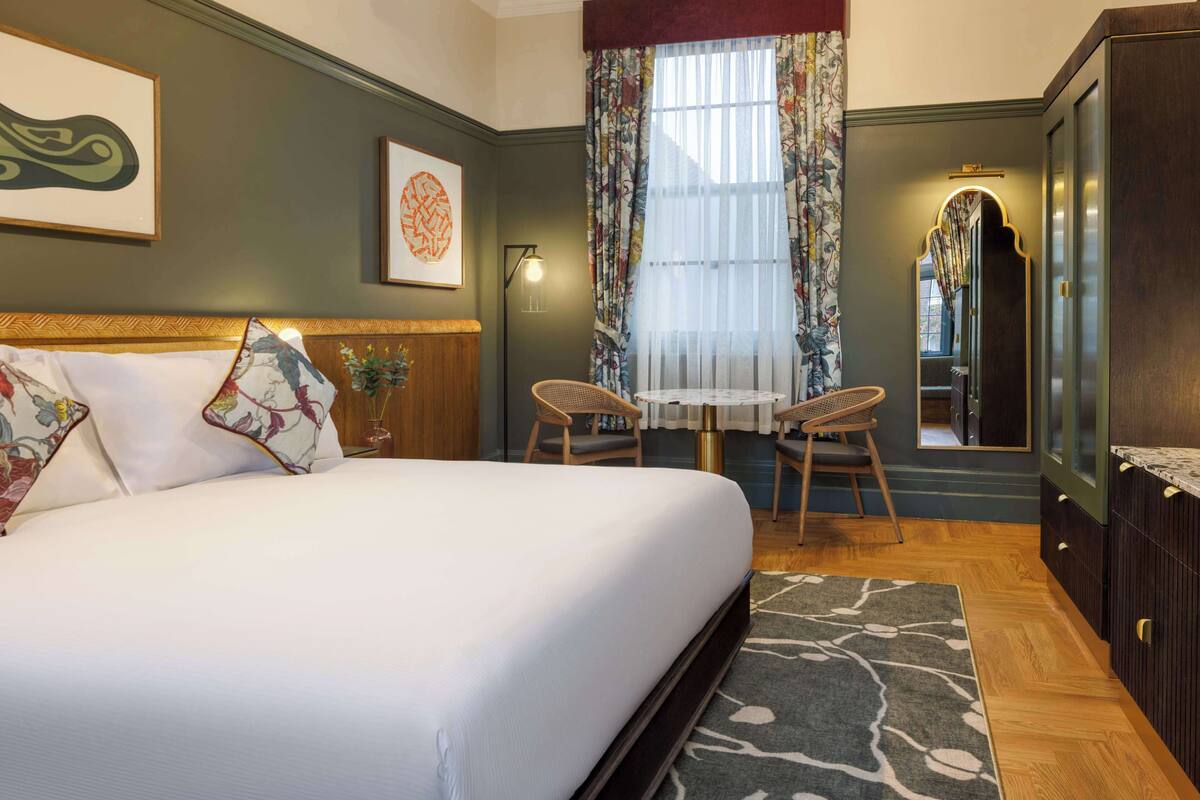 A bedroom with a large bed, a small table with two chairs, a mirror, and a wardrobe.