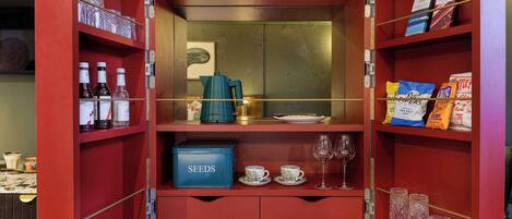 A red storage unit with shelves containing bottles, a blue box labeled 'SEEDS', and two cups on the lower shelf.