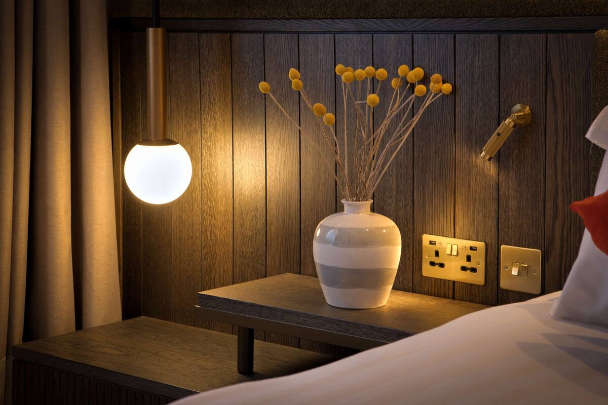 A bedside table with a vase of dried flowers, a wall-mounted light, and a power outlet.