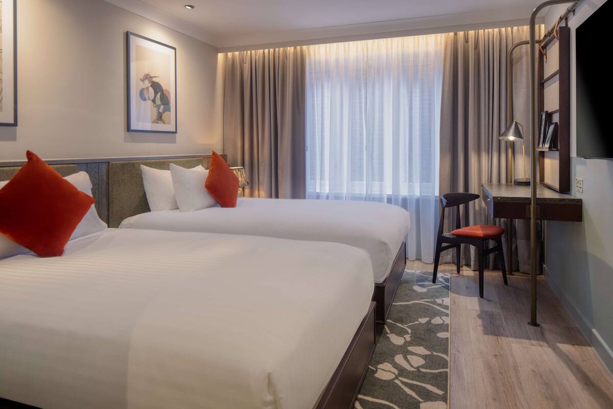 A hotel room with two beds, a desk, a chair, a TV, and a window with curtains.