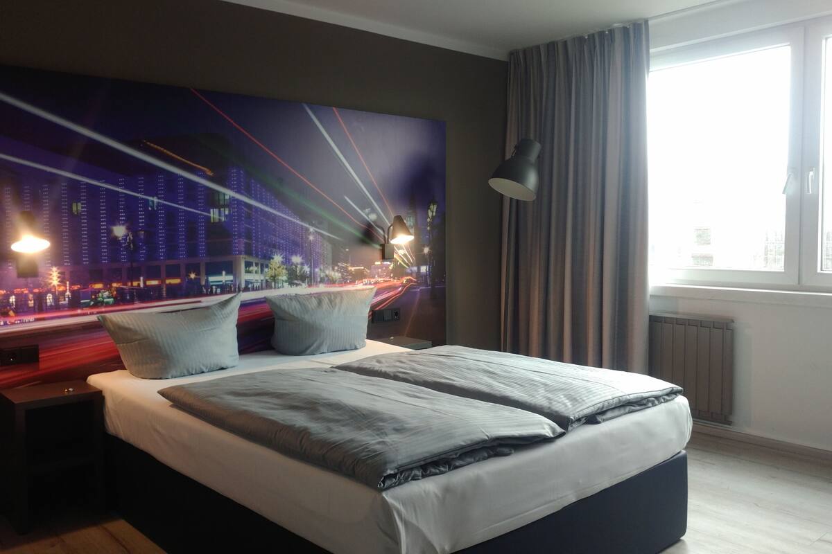 A modern hotel room with a large bed, a night cityscape mural, a desk with a lamp, and a window with curtains.