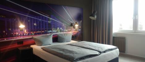 A modern hotel room with a large bed, a night cityscape mural, a desk with a lamp, and a window with curtains.