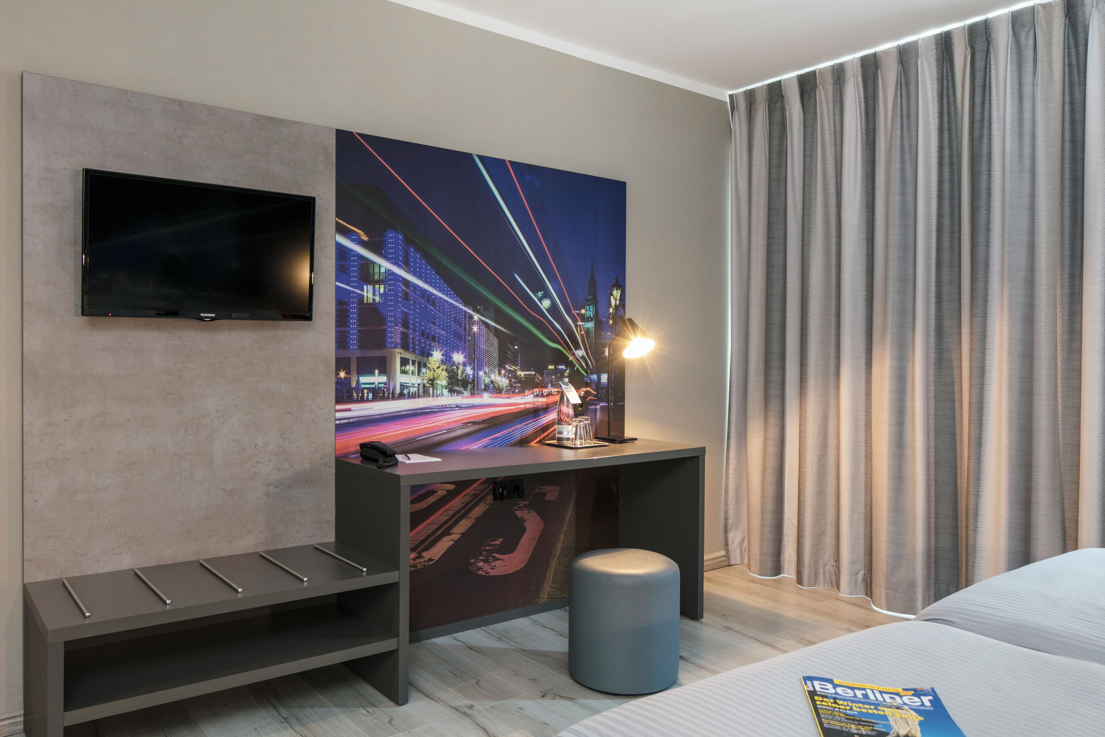 Een moderne hotelkamer met een grote flatscreen televisie aan de muur, een bureau met een tijdschrift, een bankje en een groot raam met gordijnen.