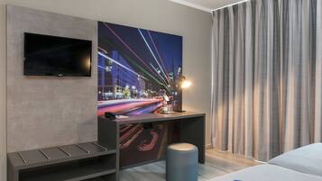 Een moderne hotelkamer met een grote flatscreen televisie aan de muur, een bureau met een tijdschrift, een bankje en een groot raam met gordijnen.