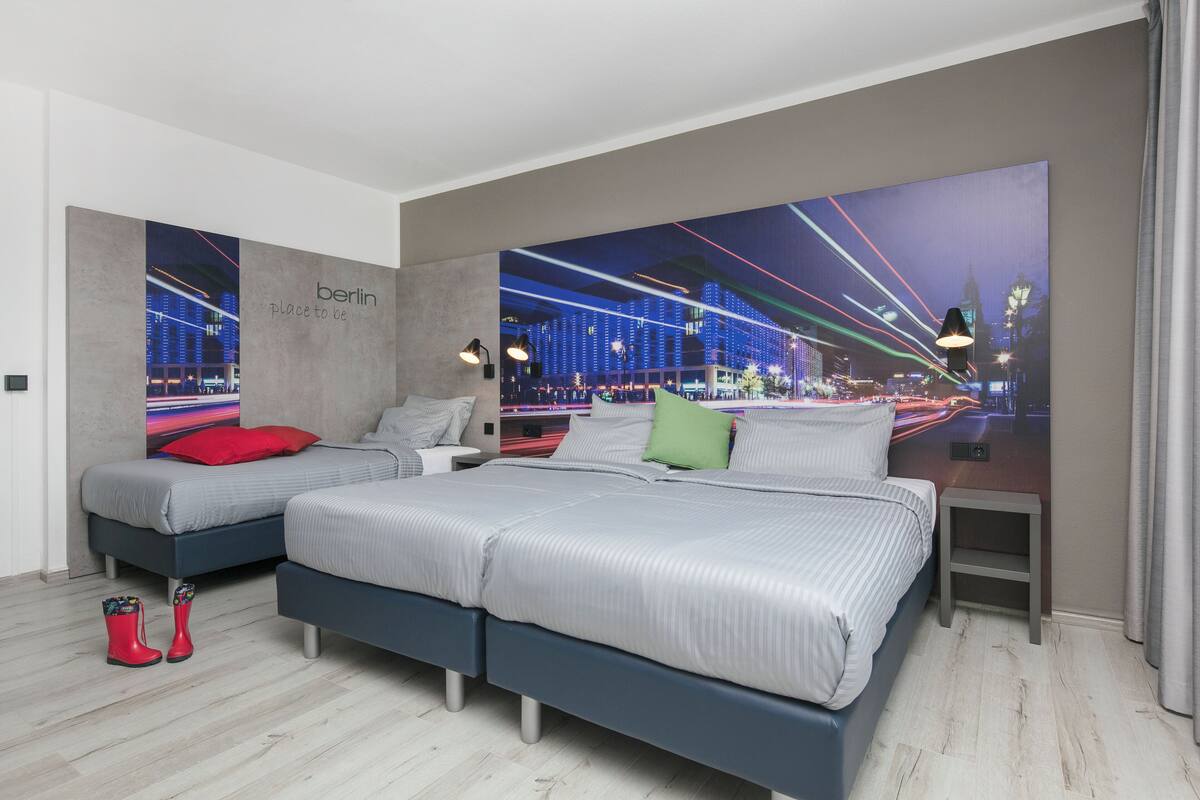 A modern bedroom with a large bed, two bedside tables, and a nightstand. There is a cityscape mural on the wall, a red pillow, and a pair of red slippers on the floor.