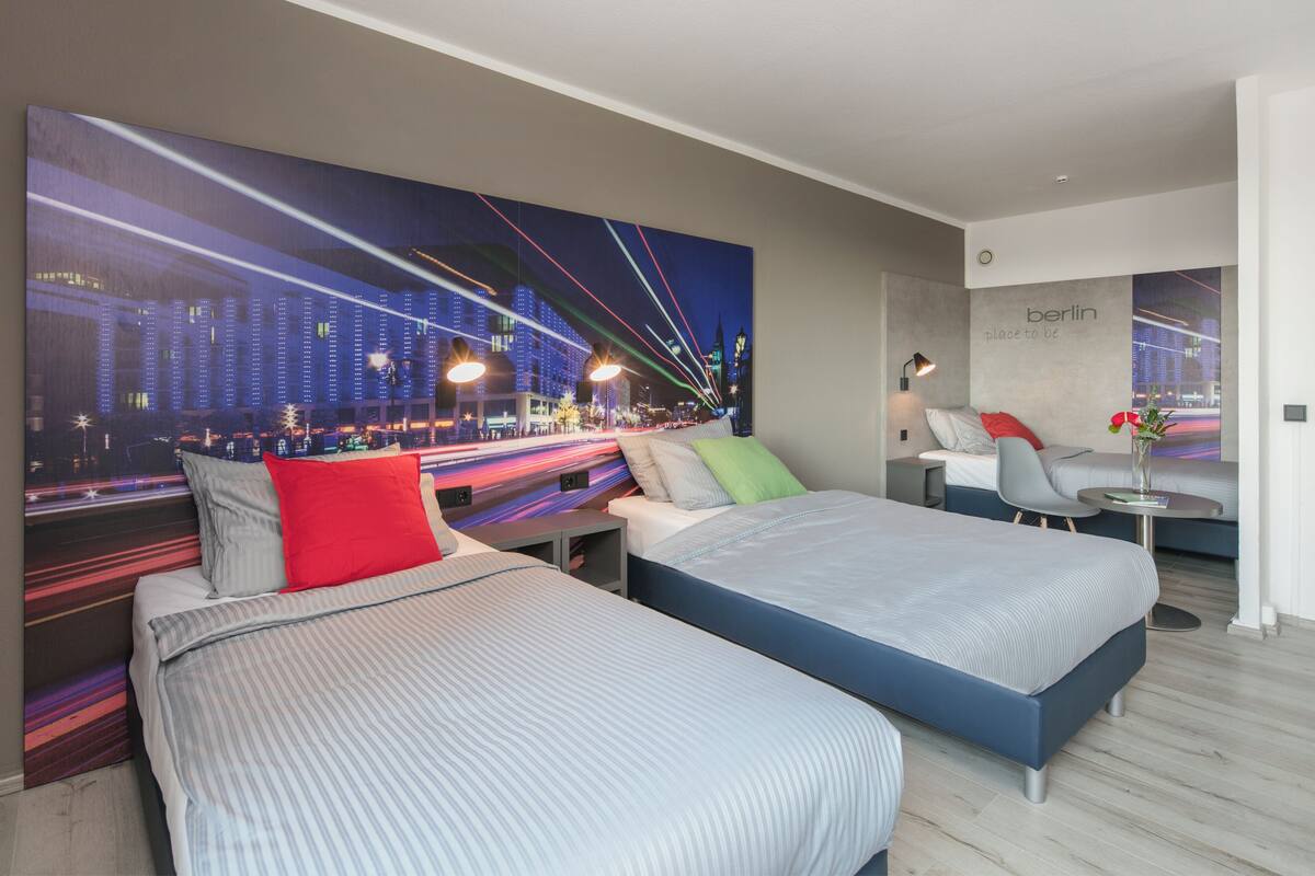 A hotel room with two beds, a large cityscape mural, a small table with a vase, and a sign that says 'berlin place to be'.