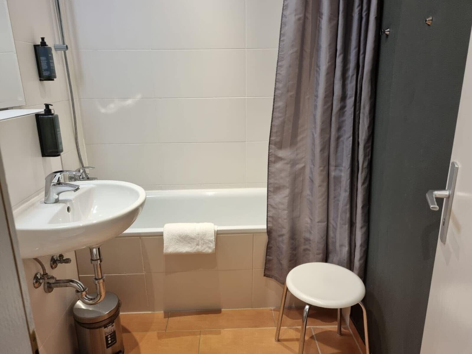 A bathroom with a white sink, a bathtub, a shower curtain, and a small stool.