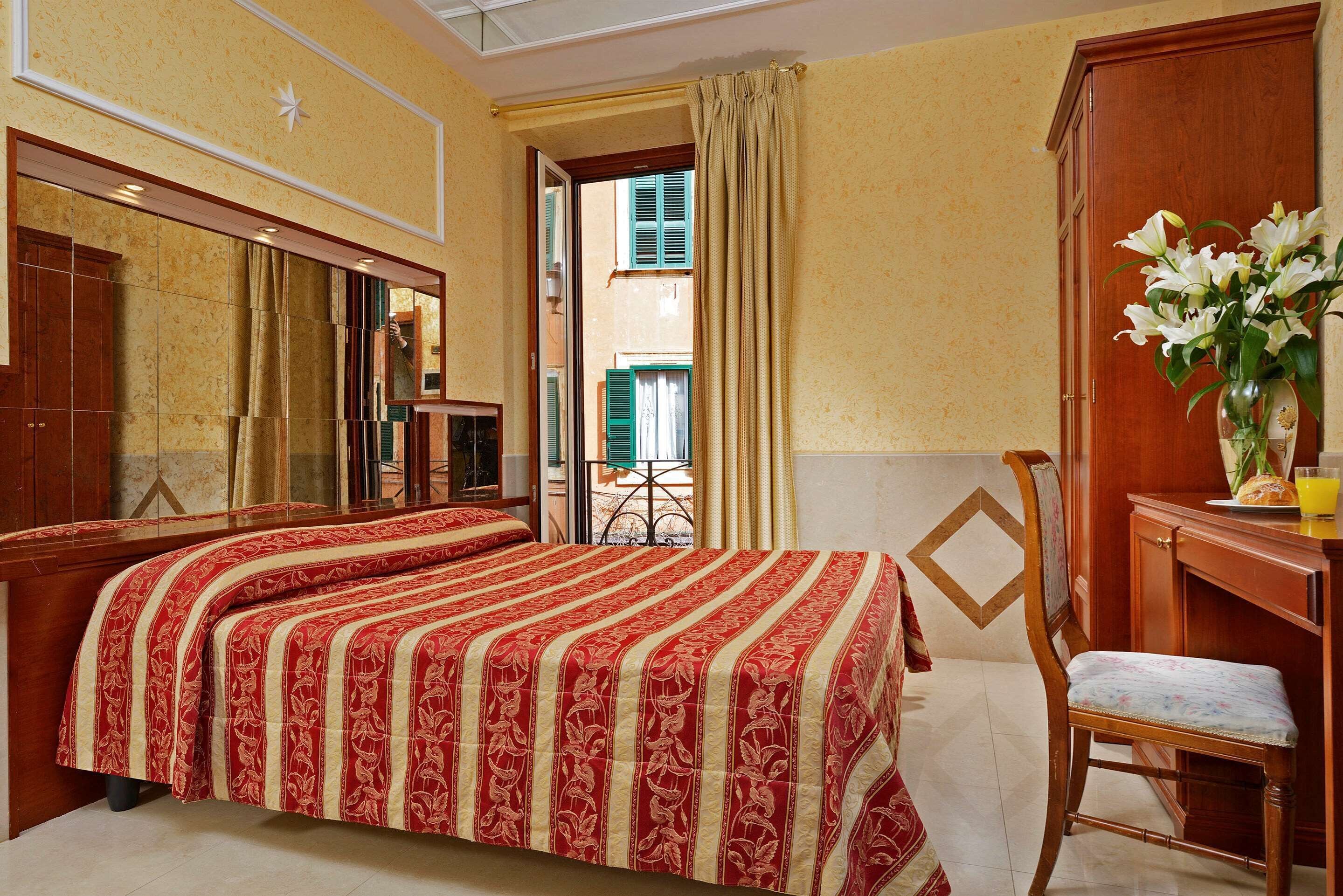 A hotel room with a large bed, a wooden chair, a desk with a vase of flowers, and a view of the street through an open door.