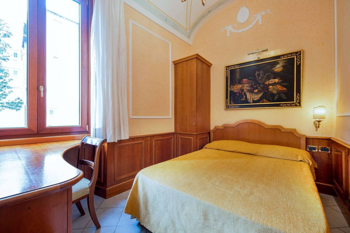 A hotel room with a wooden bed, a desk, and a chair. There is a window with curtains, a painting on the wall, and a light fixture.