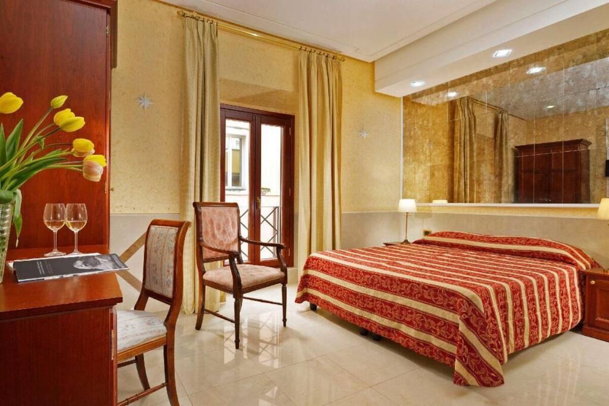 A hotel room with a bed, a desk with a vase of flowers, a chair, and a window with curtains.