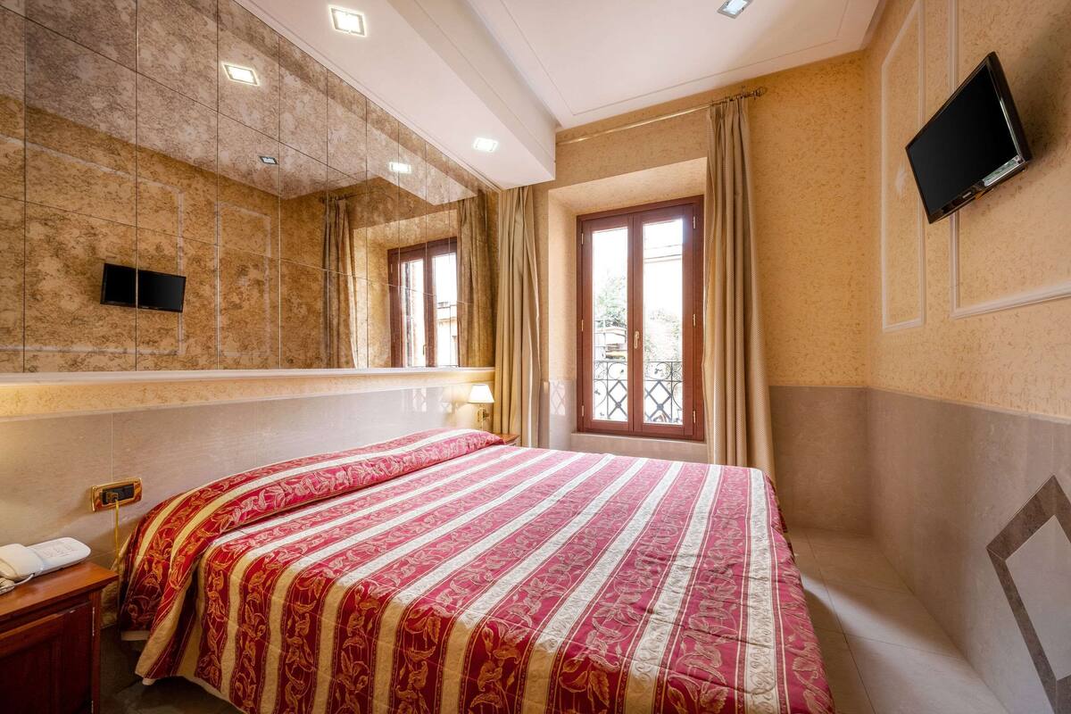 A hotel room with a large bed, a TV mounted on the wall, and a window with curtains.