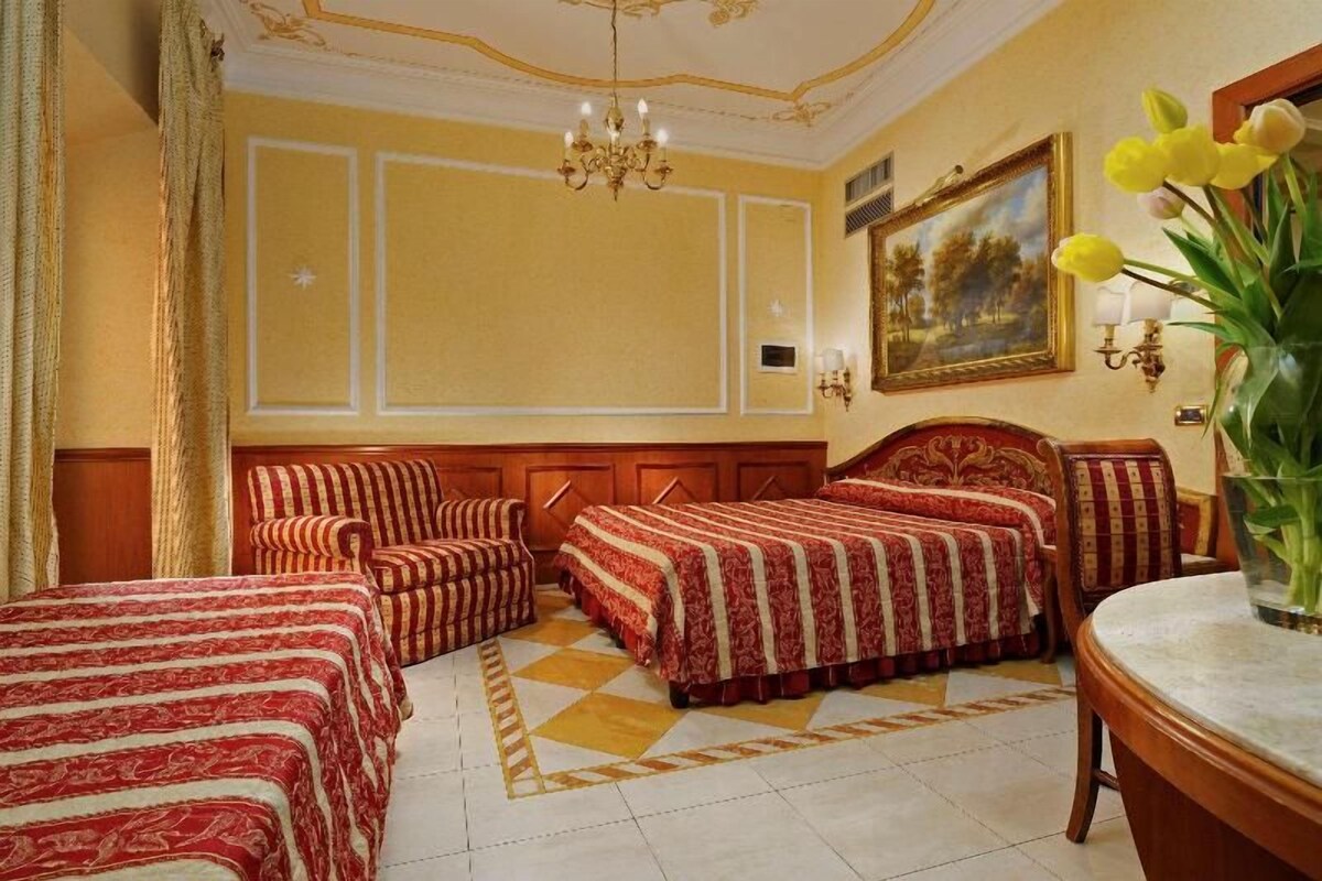 A hotel room with three beds, a chandelier, a painting, and a vase with flowers.