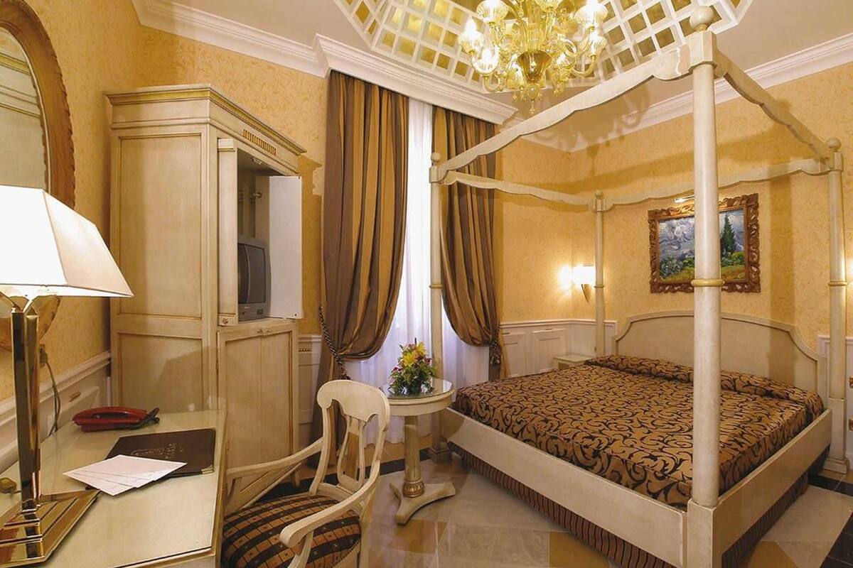 A four-poster bed, a desk with a lamp, a chair, a television, and a painting on the wall.