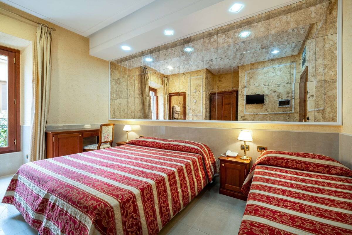A hotel room with two beds, a large mirror, a wooden desk, and a window with curtains.