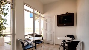 A room with a TV mounted on the wall, two tables with chairs, a door, and a large window with a view of the beach. - Motel 6 Jennings, LA (Jennings)