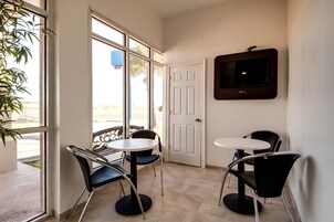 Una habitación con un televisor en la pared, dos mesas con sillas, una puerta y un ventanal con vistas a la playa.