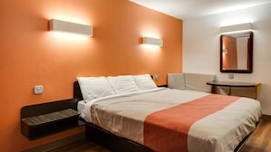 A hotel room with a bed, a bedside table, a chair, a small table, and a wall-mounted mirror. - Motel 6 Jennings, LA (Jennings)