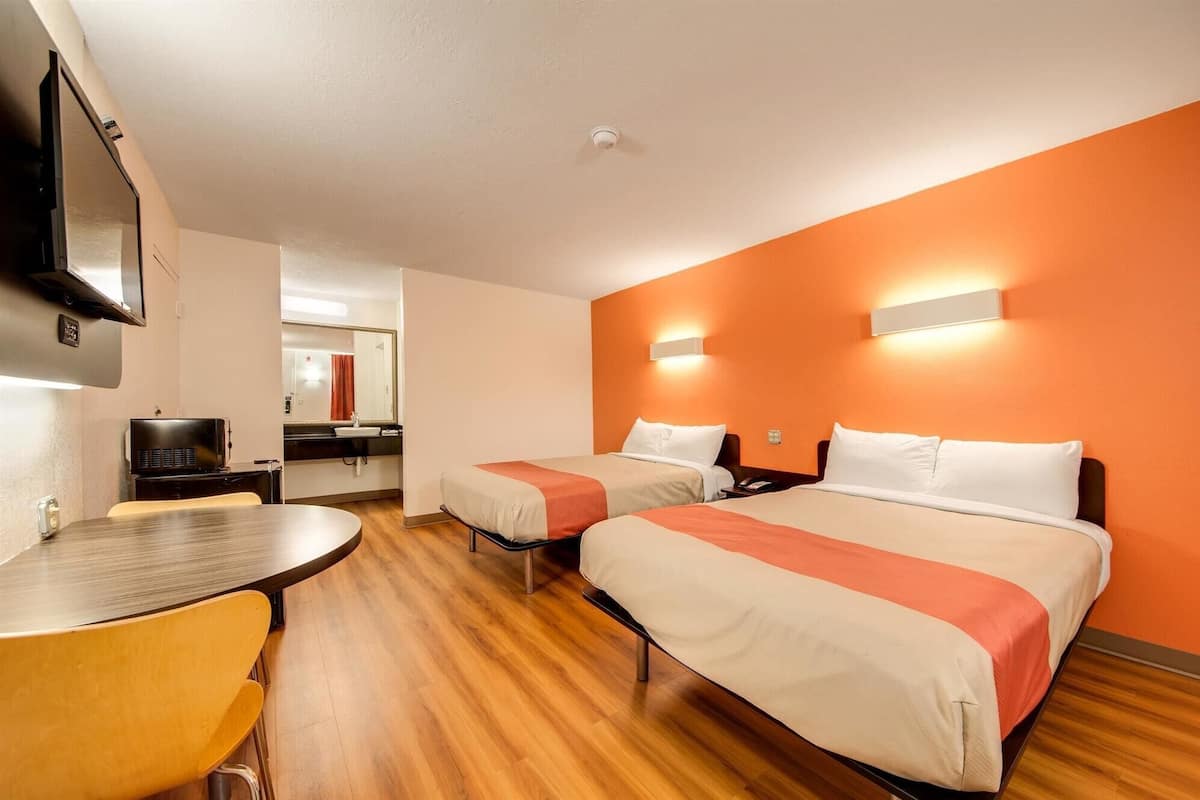 Habitación de hotel con dos camas, un escritorio, una silla, televisión y baño a la vista.