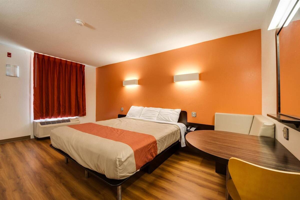 Habitación de hotel con cama, escritorio y silla. Hay dos luces fijadas en la pared sobre la cama y una ventana con cortinas rojas.