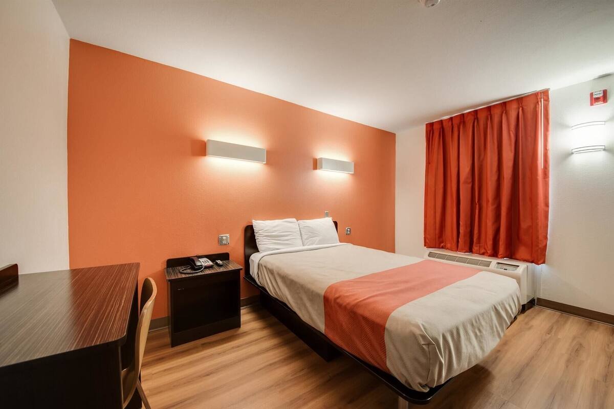 Habitación de hotel con cama, escritorio, silla, mesita de noche con teléfono y ventana con cortinas.