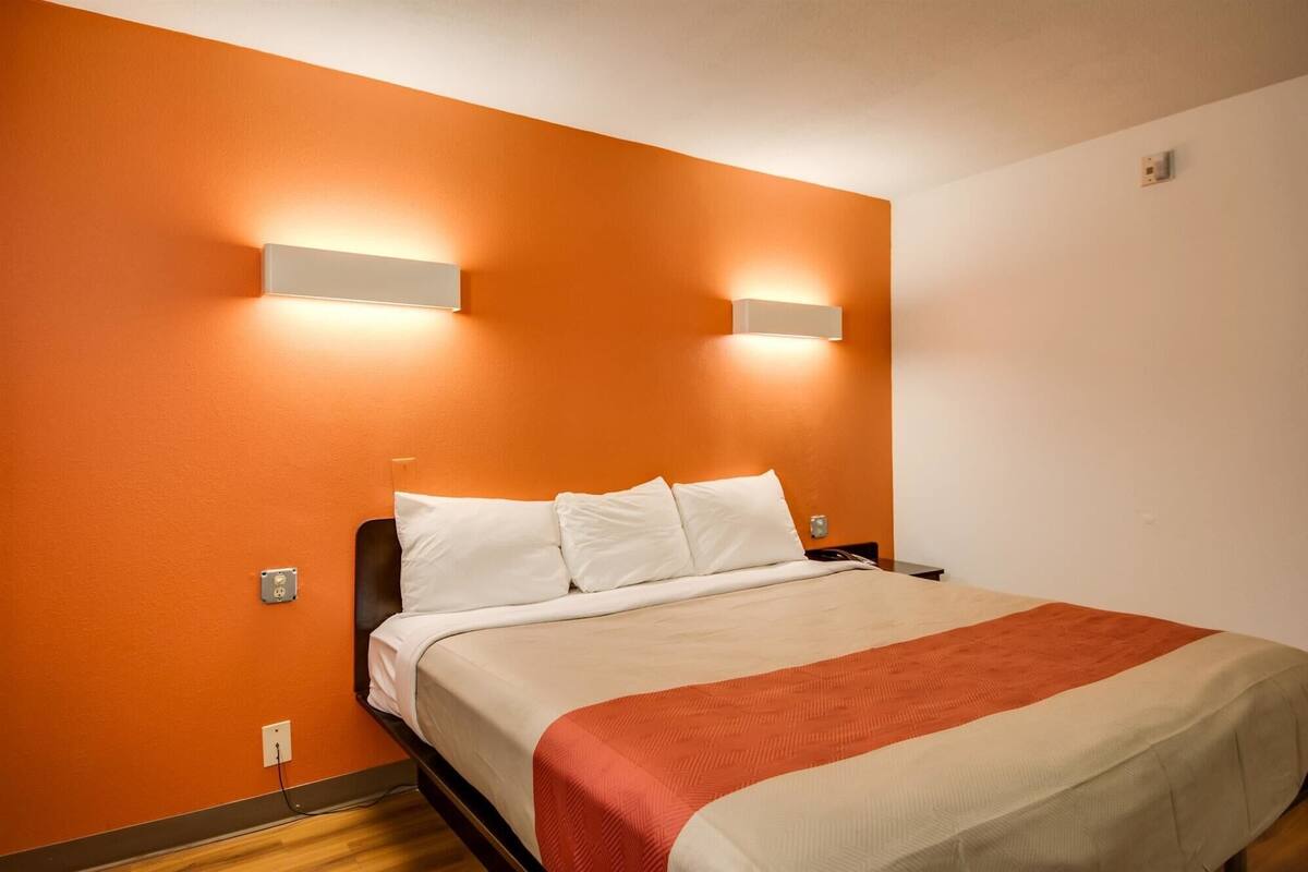 Una habitación de hotel con pared naranja, cama con ropa de cama blanca y roja, y dos lámparas fijadas a la pared.