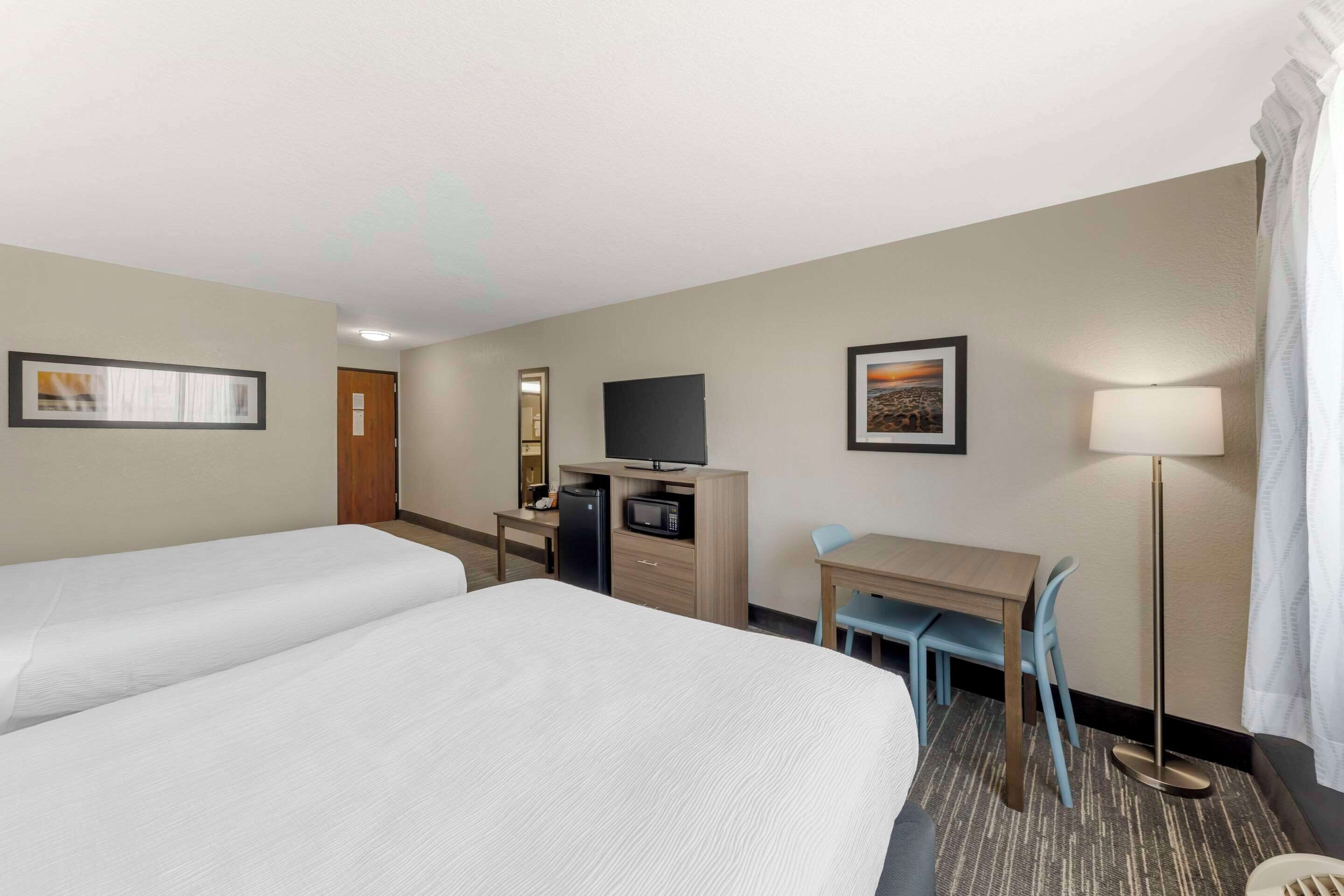 A hotel room with two beds, a desk with a computer and a microwave, a TV, a lamp, and a chair.