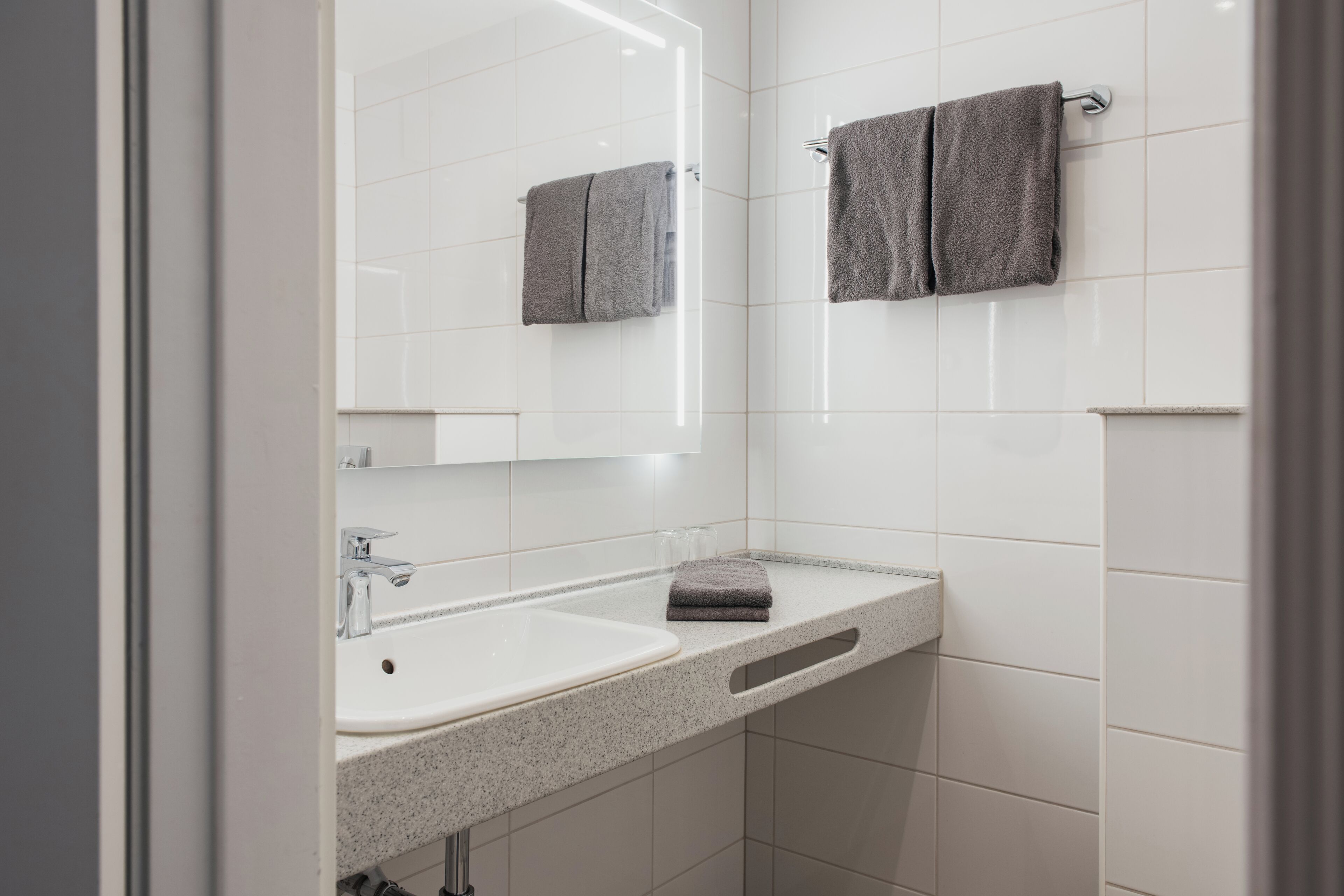 Un baño con un lavamanos blanco, un espejo y dos toallas colgadas en la pared.