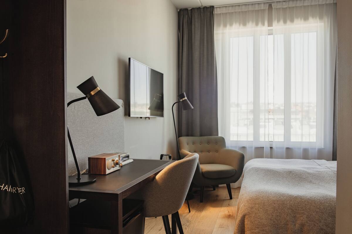 Hotellihuone, jossa on työpöytä, tuoli, sänky, televisio ja suuri ikkuna, jossa on verhot.
