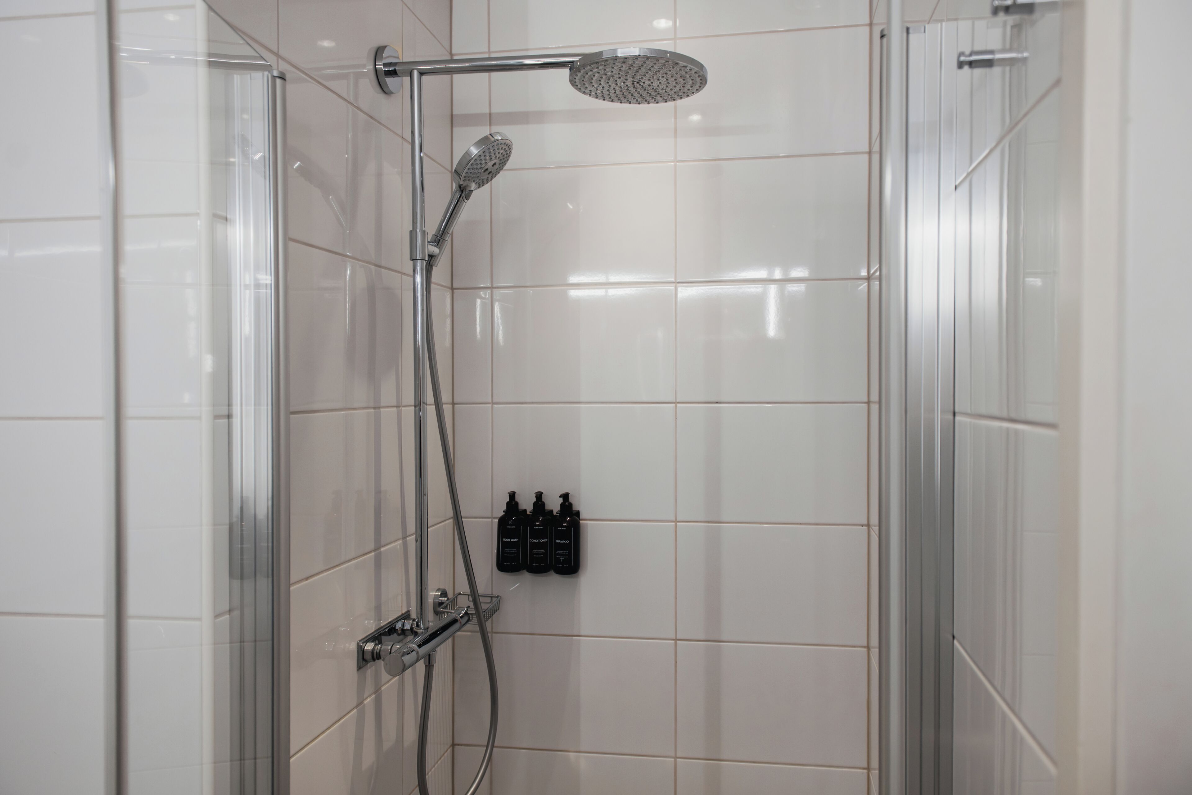 Una ducha con cabezal de ducha manual y otro tipo lluvia empotrado en la pared, mampara de vidrio y tres botellas negras en la pared.