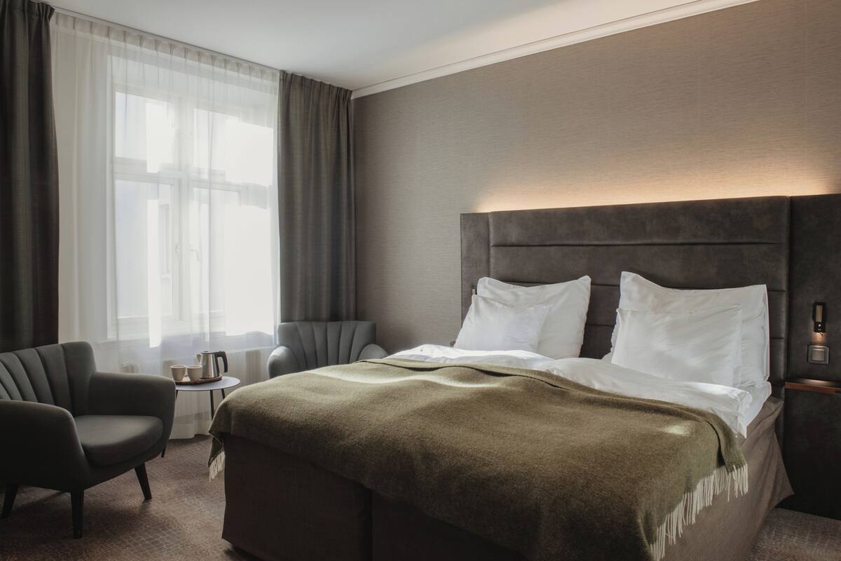 Hotellihuone, jossa on sänky, tuoli, pieni pöytä kahvinkeittimellä sekä ikkuna, jossa on verhot.