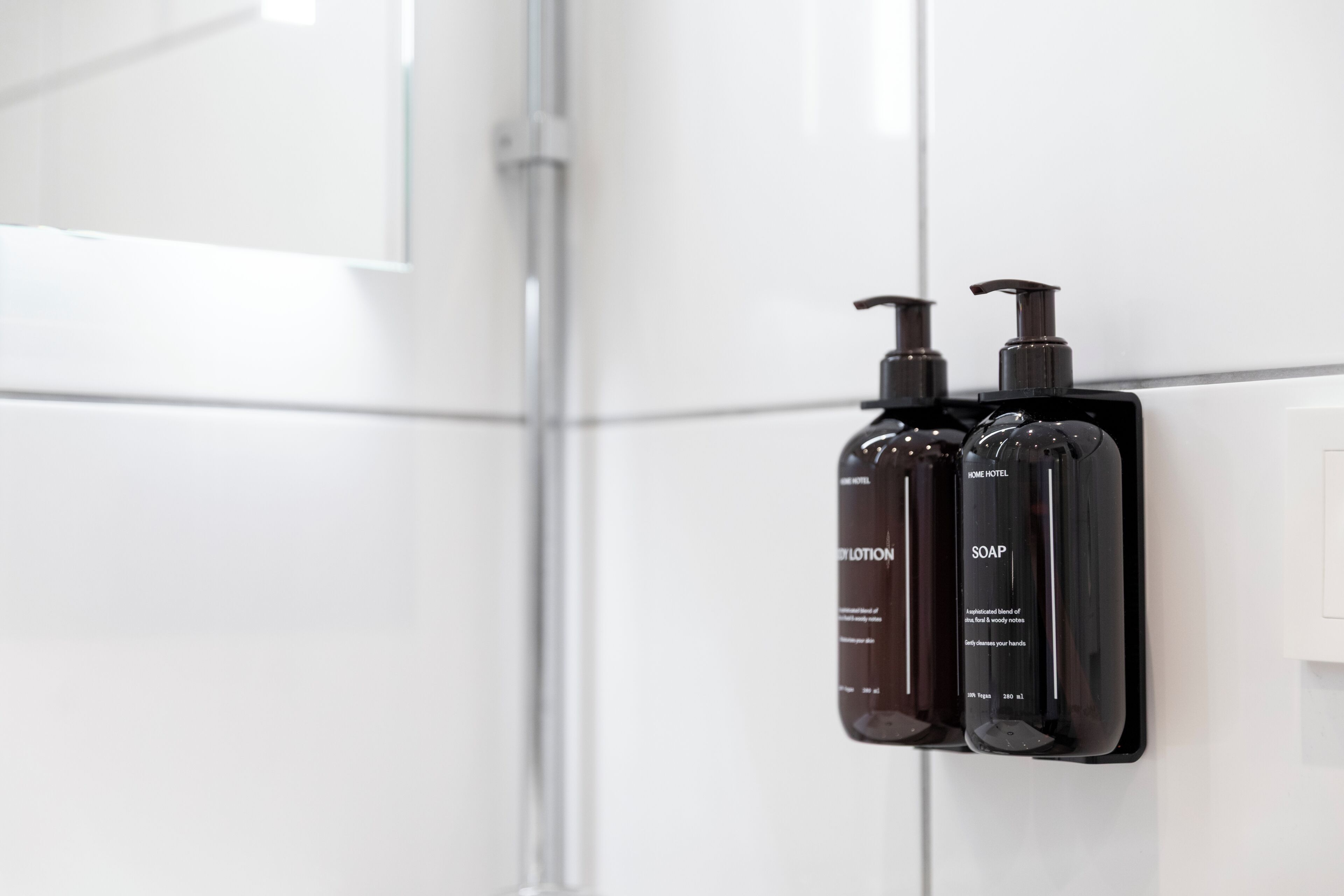Two black bottles of lotion and soap mounted on a white tiled wall.