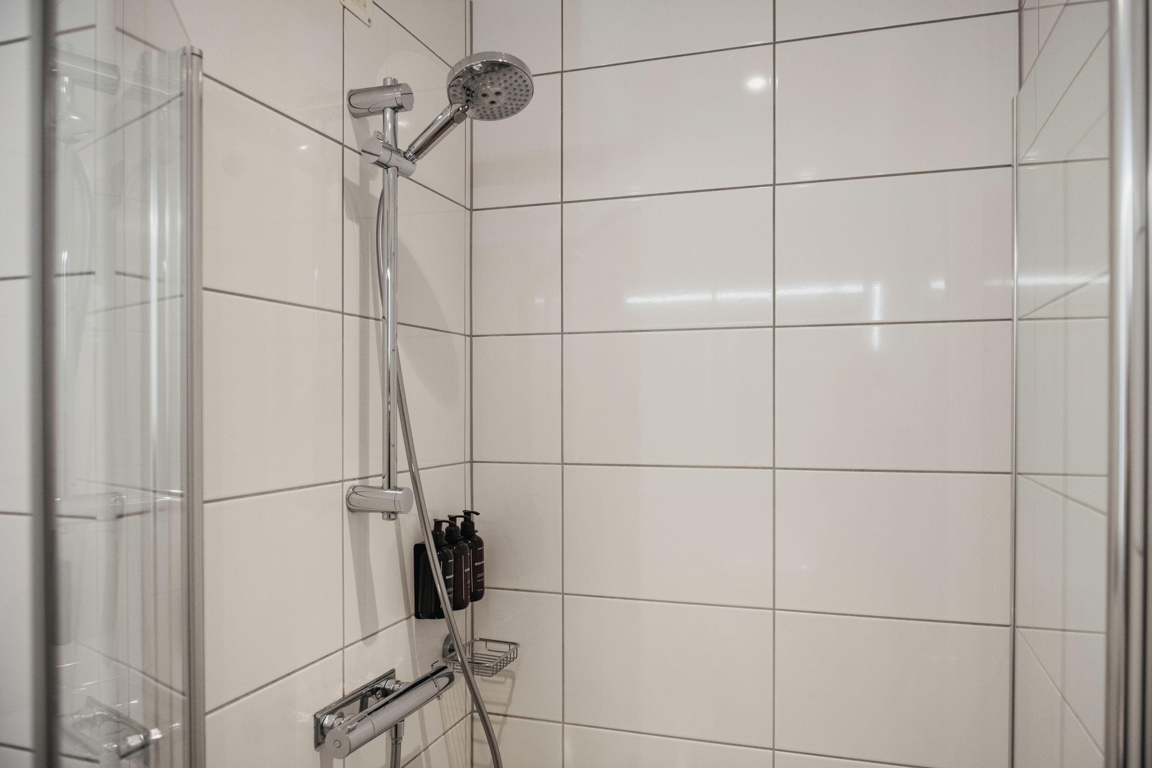 En modern dusch med ett justerbart duschhuvud, en väggmonterad kontrollpanel och en liten hylla för toalettartiklar.