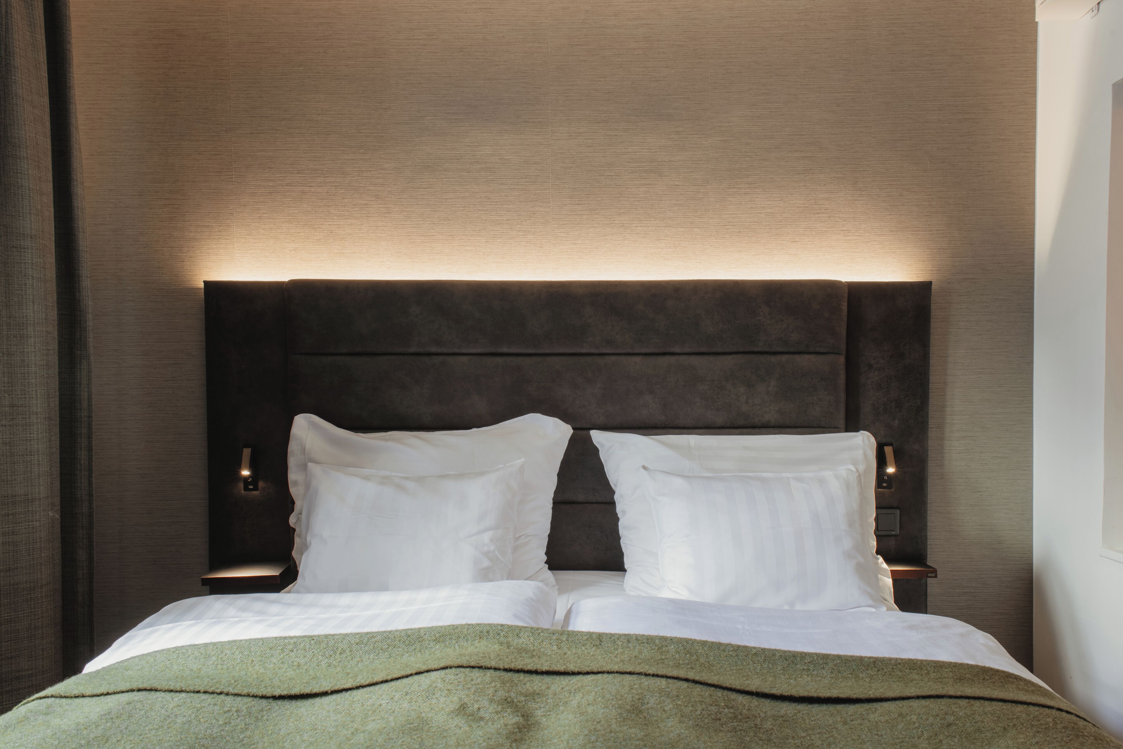 Una cama bien tendida con sábanas blancas y un plaid verde, cabecero oscuro y lámparas de noche.