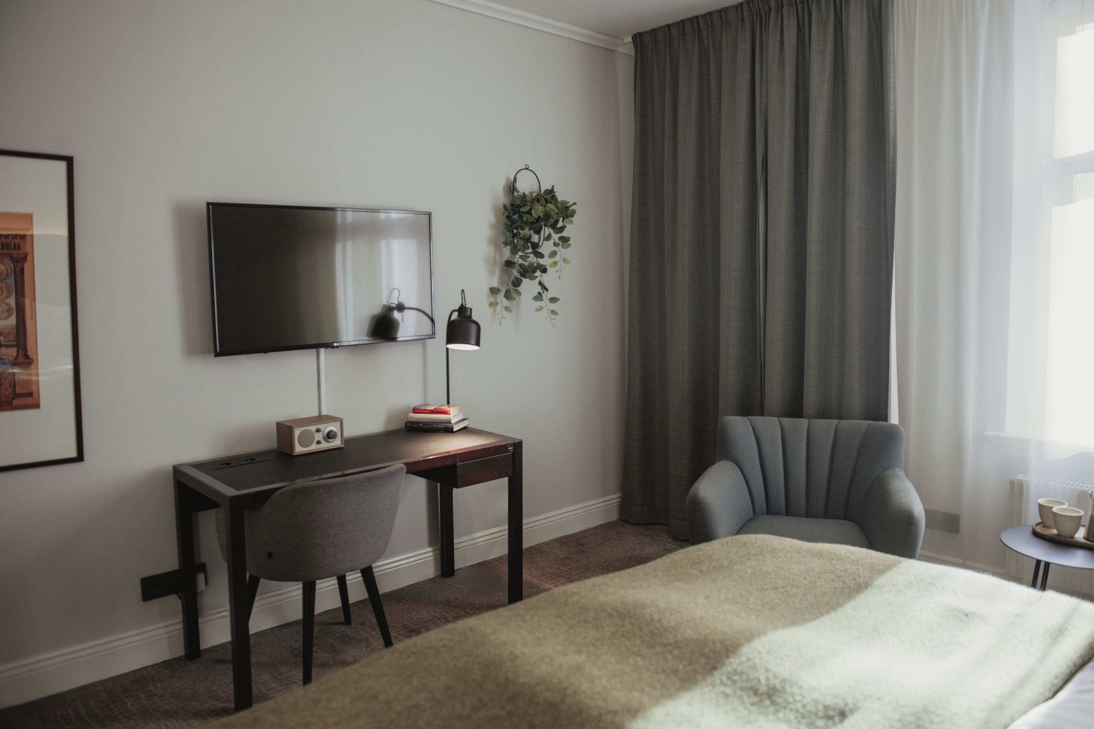 Ein Hotelzimmer mit Bett, Schreibtisch mit Stuhl, Fernseher, einem kleinen Tisch mit Lampe und einer Pflanze.