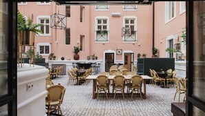 An outdoor dining area with wicker chairs and tables, potted plants, and a cobblestone floor.