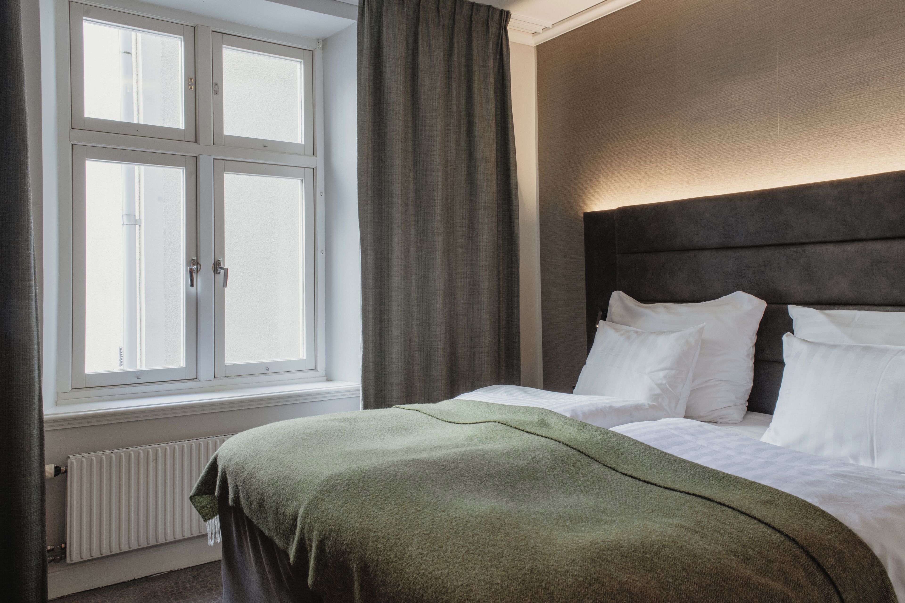 A hotel room with a bed, a window with curtains, a radiator, and a headboard.