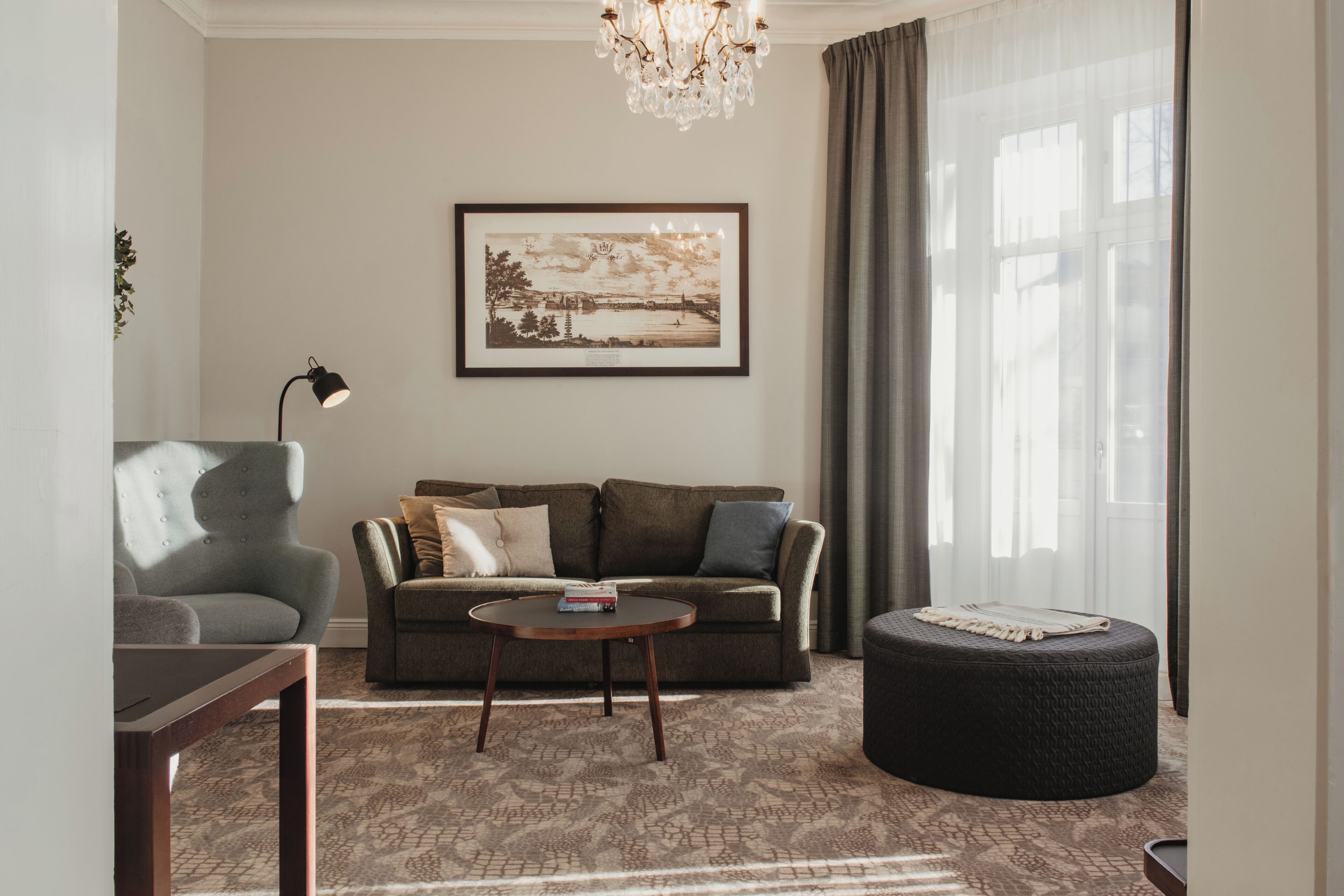 A living room with a sofa, a round coffee table, a chair, a round ottoman, a framed picture on the wall, and a chandelier.