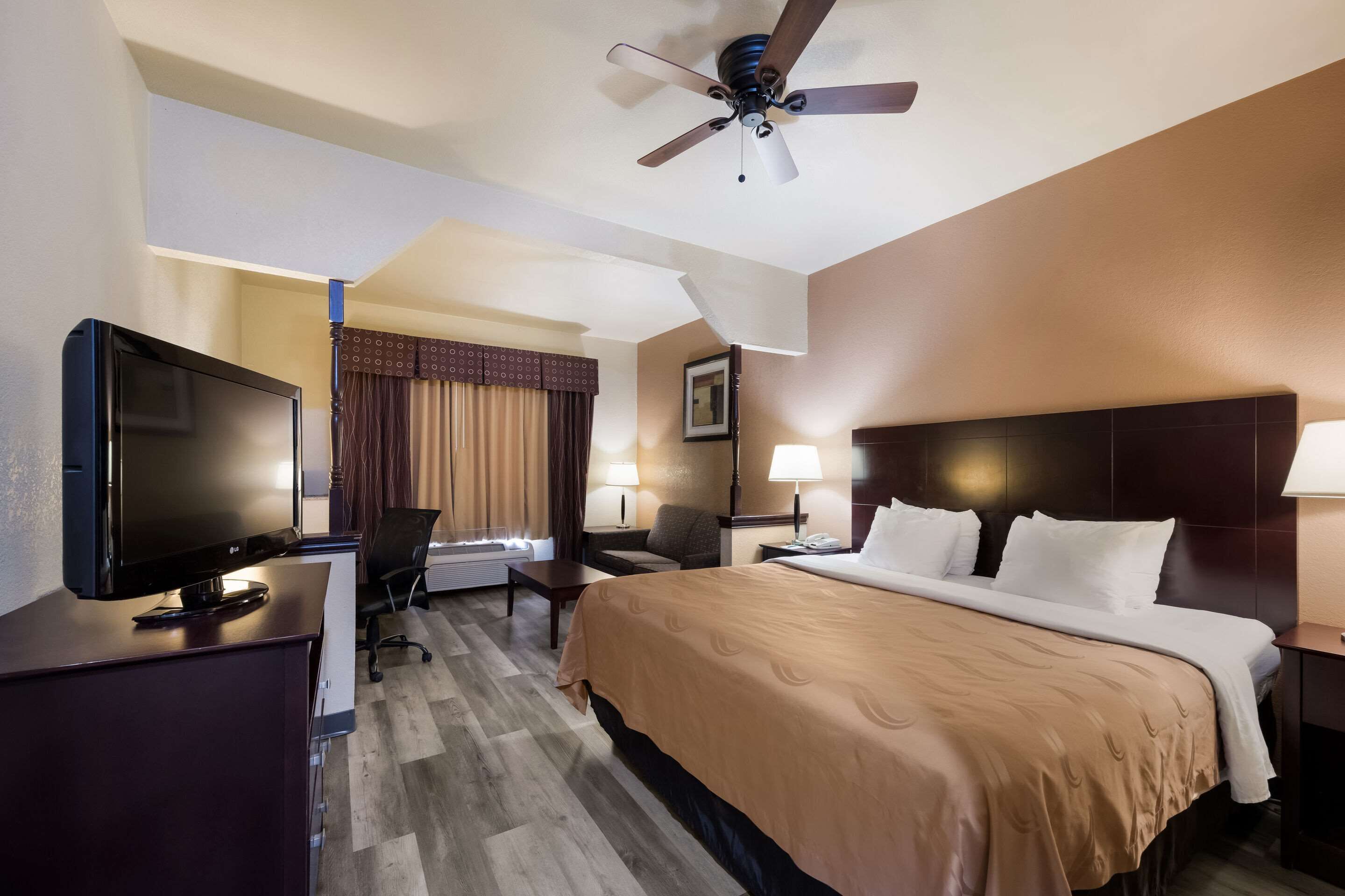 A hotel room with a bed, a desk with a TV, a chair, a desk lamp, a nightstand, and a ceiling fan.