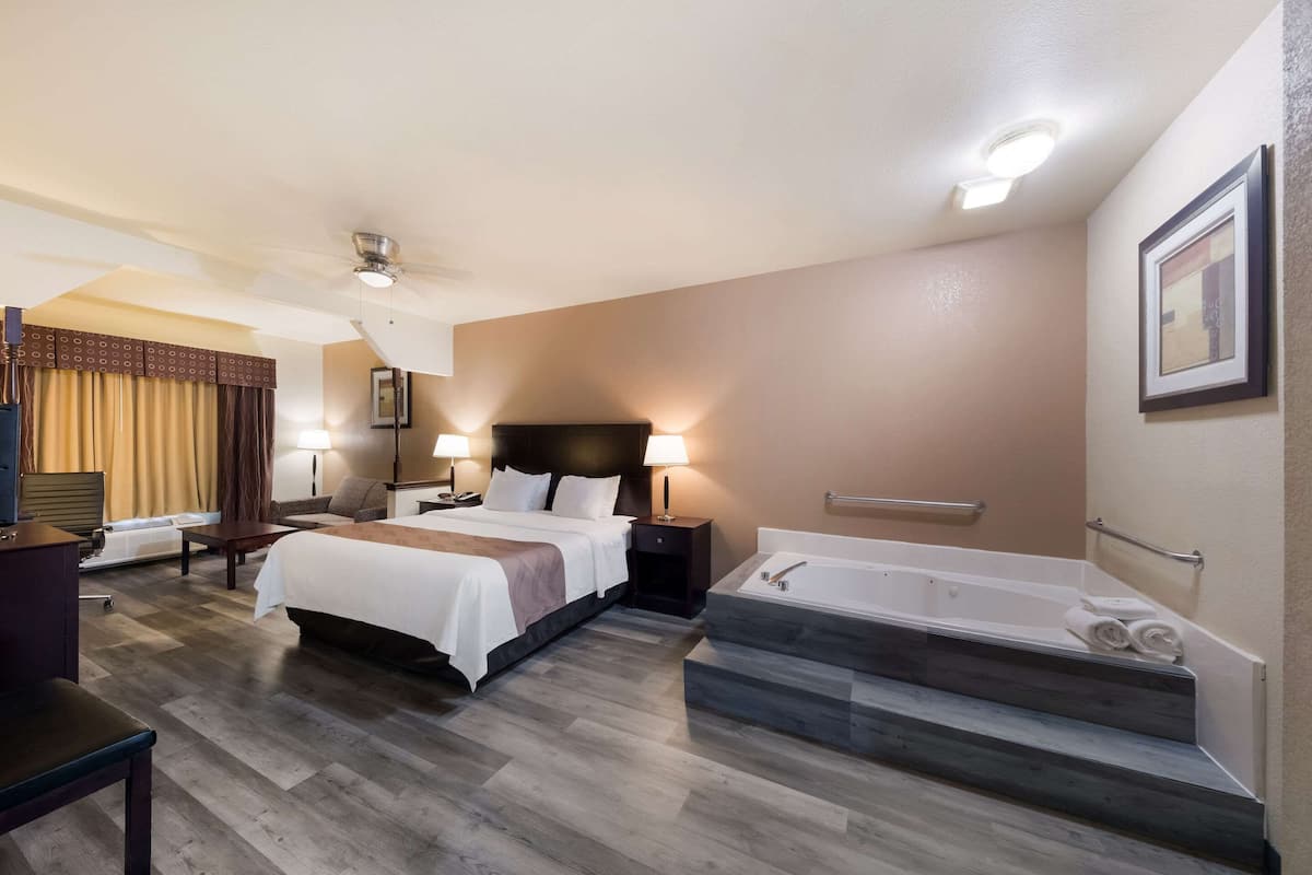 A hotel room with a bed, bedside tables, a desk, a chair, a large window with curtains, a bathtub, and a shower area.