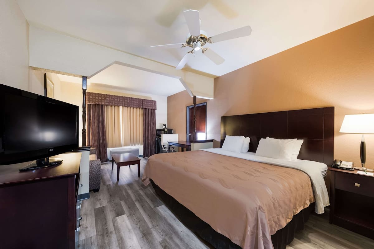 A hotel room with a bed, a TV, a desk, a chair, a ceiling fan, and a nightstand with a lamp.