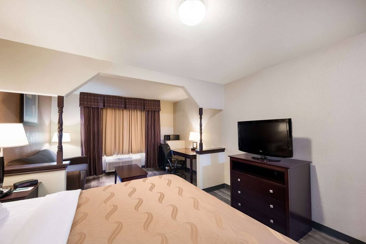 Habitación de hotel con cama, escritorio, silla, televisor y ventana con cortinas.