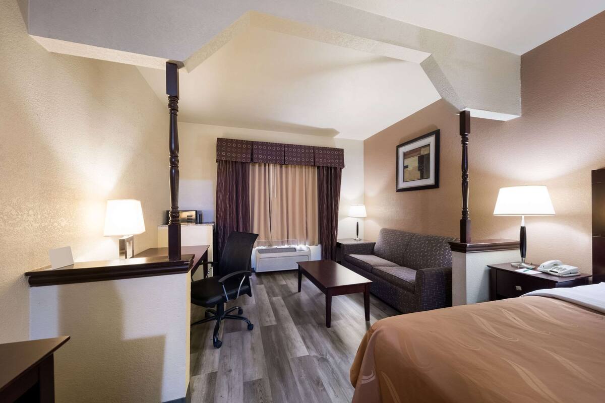 A hotel room with a desk, a chair, a sofa, a bed, a lamp, a telephone, and a picture on the wall.