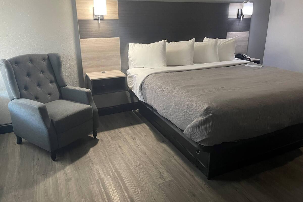 A hotel room with a bed, a chair, a nightstand, and a wall-mounted lamp.