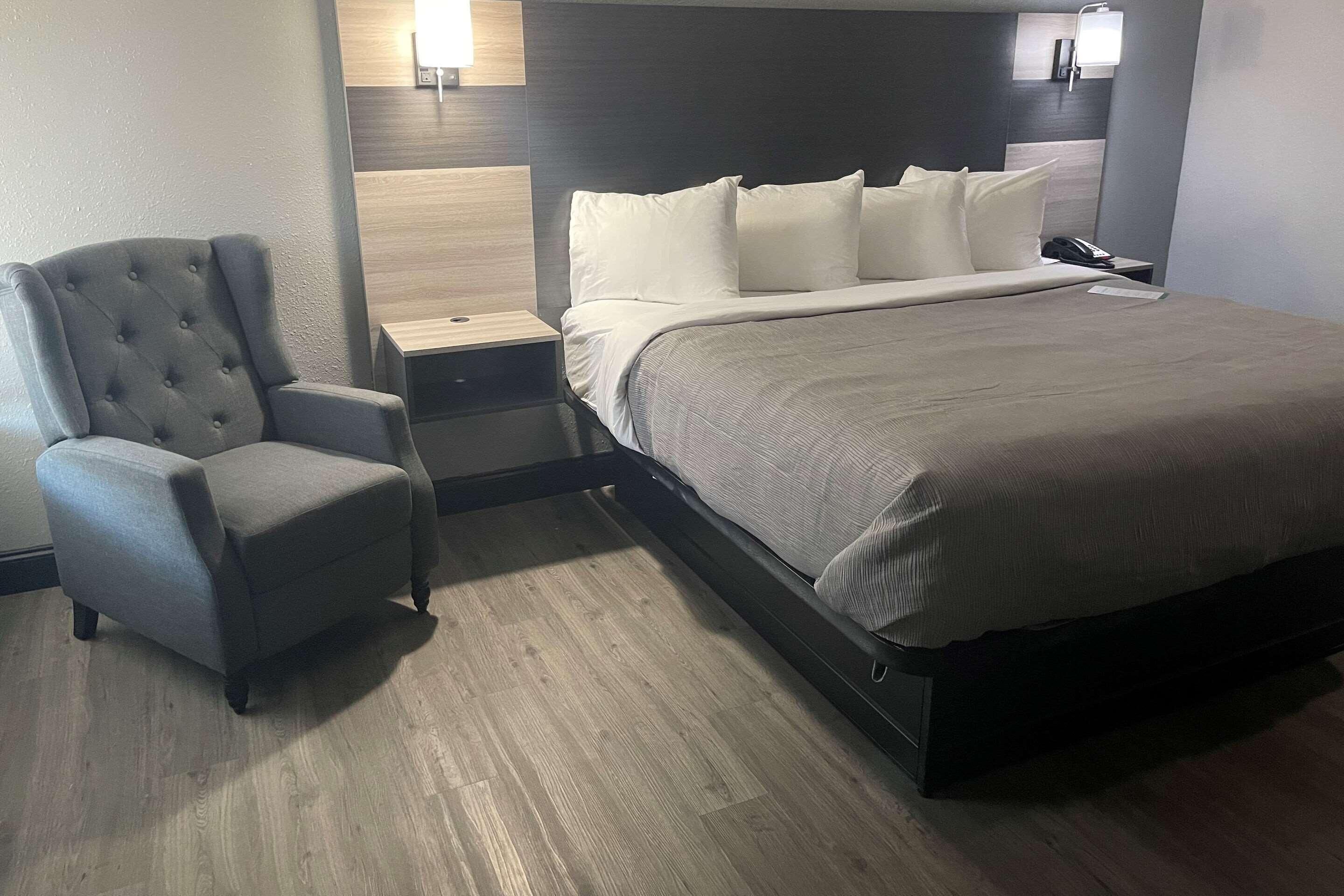 Una habitación de hotel con cama, una silla, una mesita de noche y una lámpara de pared.