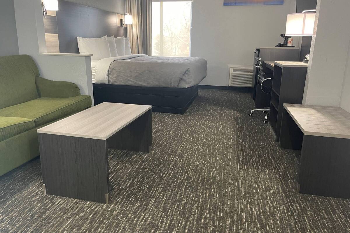 A hotel room with a bed, a green sofa, two small tables, a desk, and a window with curtains.