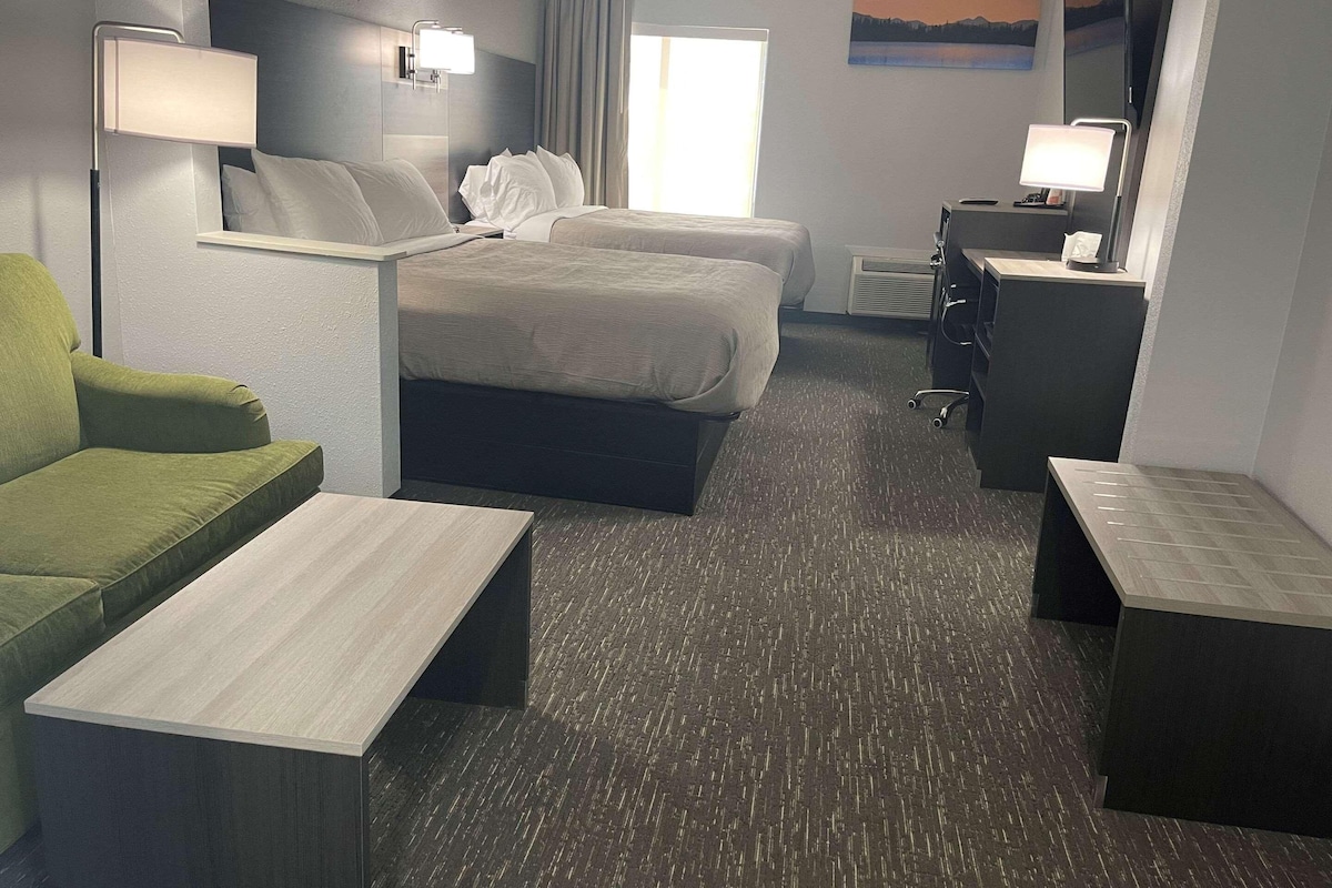 A hotel room with two beds, a desk, a chair, a small table, and a lamp.