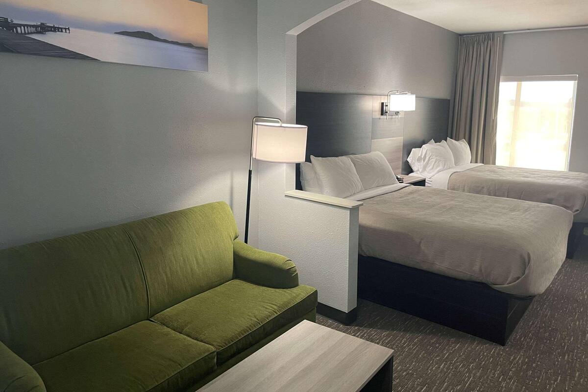 A hotel room with a green sofa, a bed with white bedding, a bedside table, a lamp, and a view of the sea.