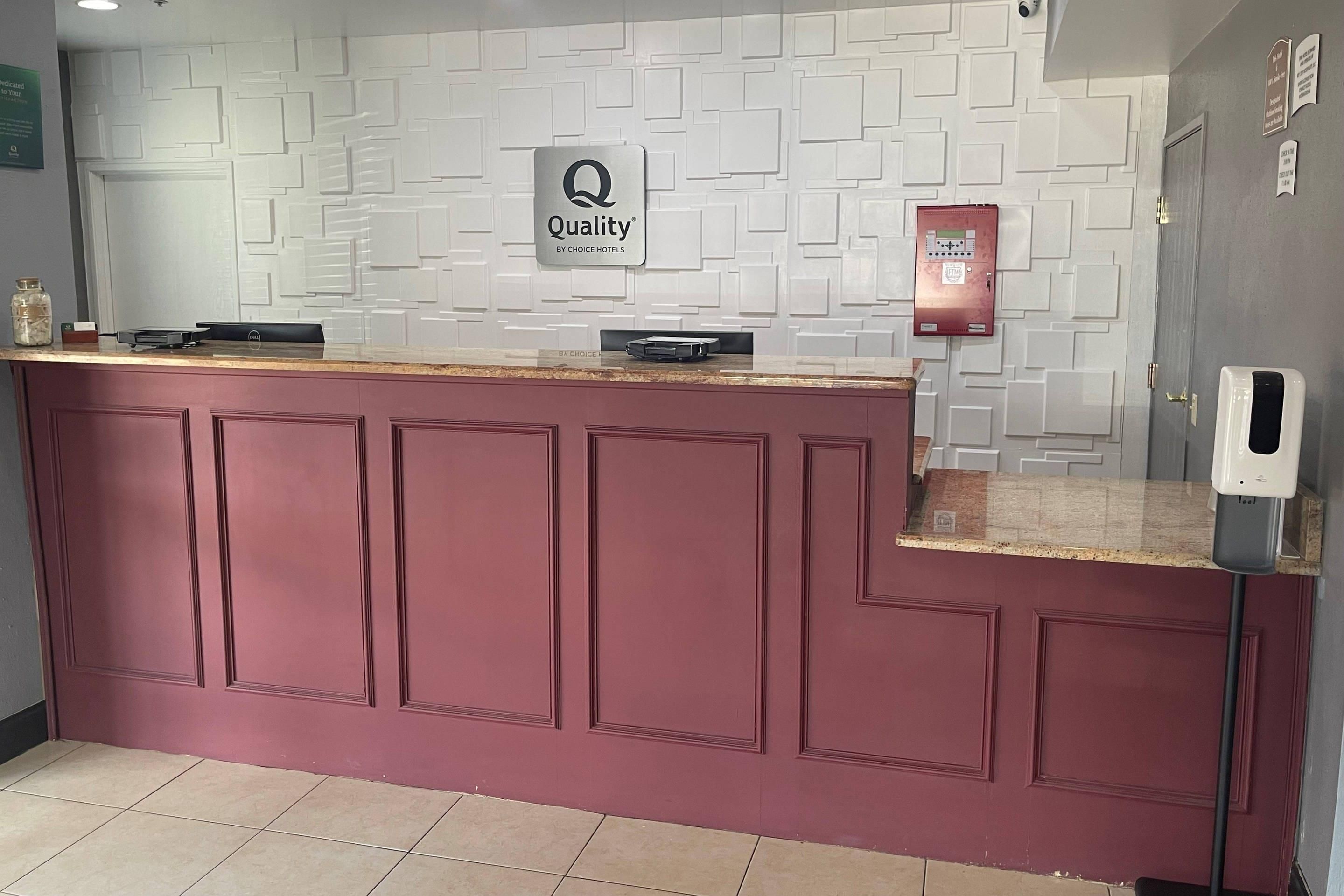 A reception desk with a 'Quality by Choice Hotels' sign, a fire alarm, and a hand sanitizer dispenser.