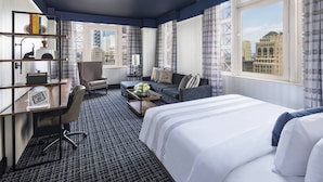 A hotel room with a large bed, a desk with a chair, a sofa, and a view of the cityscape through large windows.
